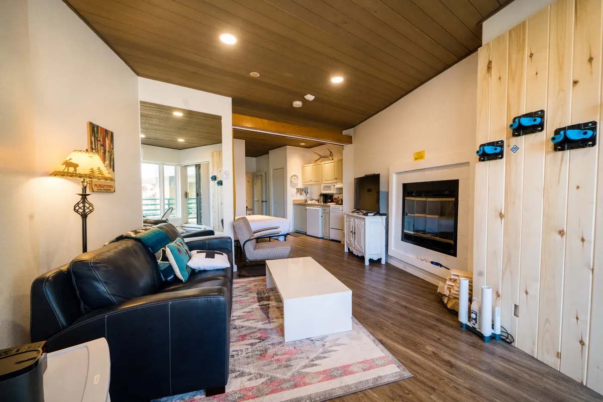 The image showcases a warm, inviting condo interior with a plush sofa, crackling fireplace, stylish coffee table, tasteful mountain-themed artwork, and a modern kitchen area. Natural light streams in, illuminating the cozy living space designed for relaxing after a day on the slopes or gathering with loved ones.