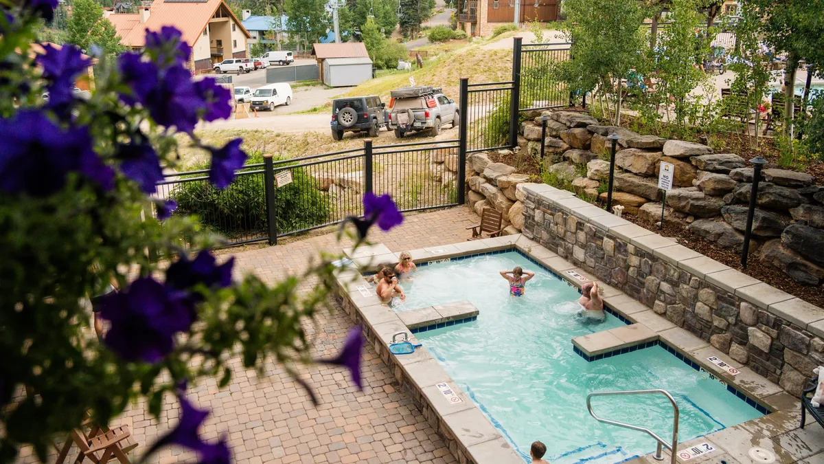 A vibrant outdoor swimming pool surrounded by flowering plants and a scenic landscape. Guests are seen enjoying the pool and relaxing in the nearby hot tub, with the cheerful atmosphere inviting for both socializing and quiet moments of relaxation under the sun.