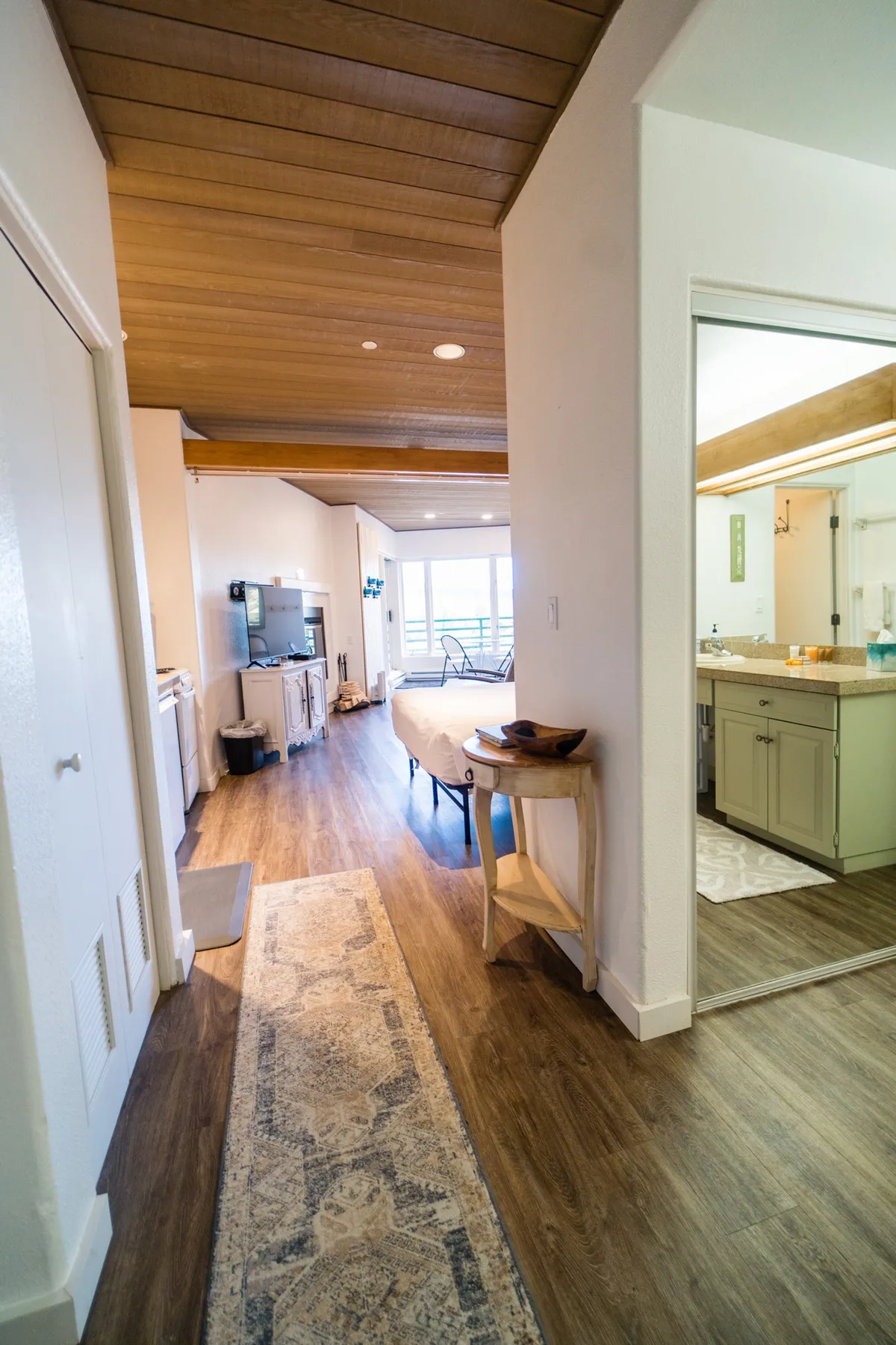 This image features an open living space with a wooden ceiling, a glimpse of the kitchen area, and a window that hints at exterior views. The decor is warm and inviting, with a decorative rug and ambient lighting that make the space feel cozy. The scene suggests opportunities to relax, gather, and enjoy both comfort and connection to the outdoors.