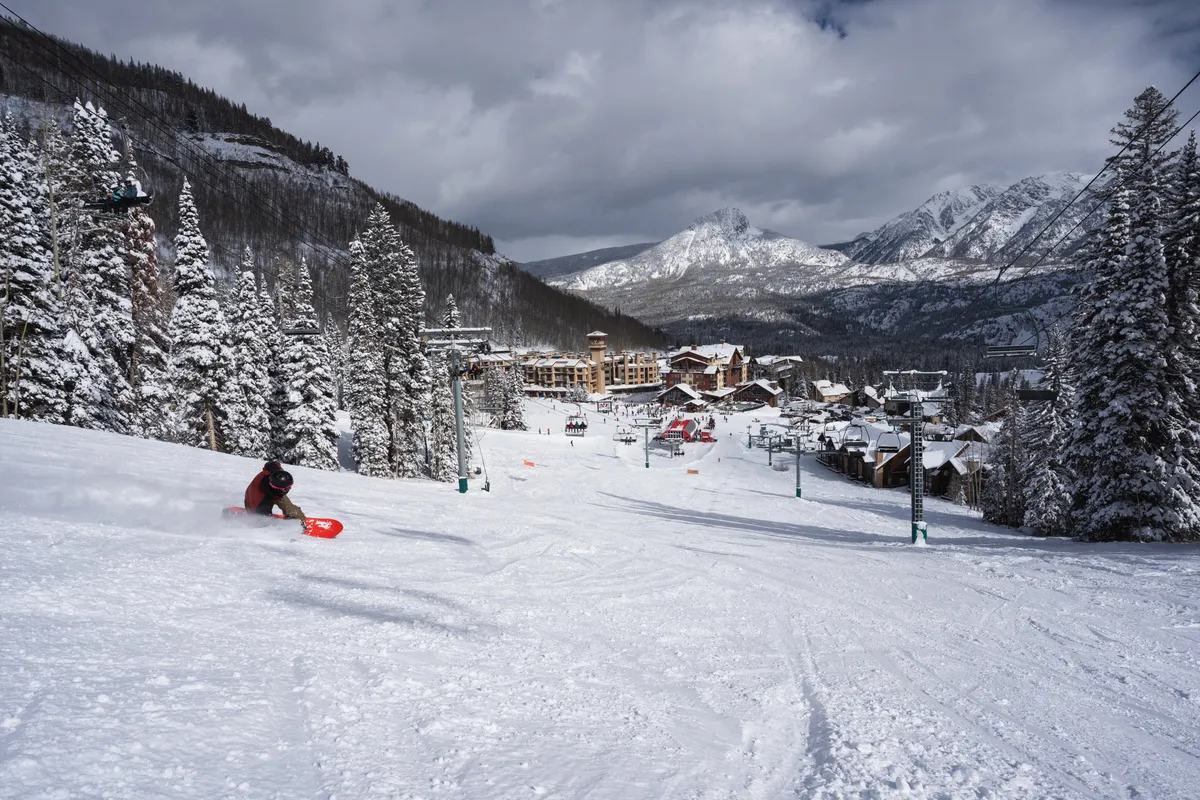 A lively exterior view showing a skier with a sled near snow-covered trees, with the ski lifts, mountains, and resort buildings in the background. The image conveys a joyful and adventurous winter atmosphere, highlighting both the excitement of skiing and the charm of the mountain setting.