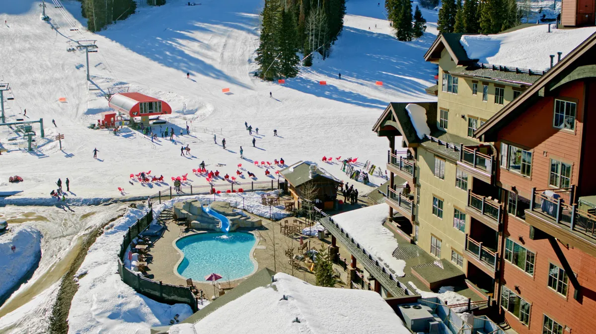 A lively exterior shot of Purgatory Village Plaza featuring snow-covered ski slopes bustling with skiers and snowboarders, a welcoming pool area, and resort buildings set against a bright, clear sky. The image captures the energetic social atmosphere and the proximity of resort amenities to the slopes.