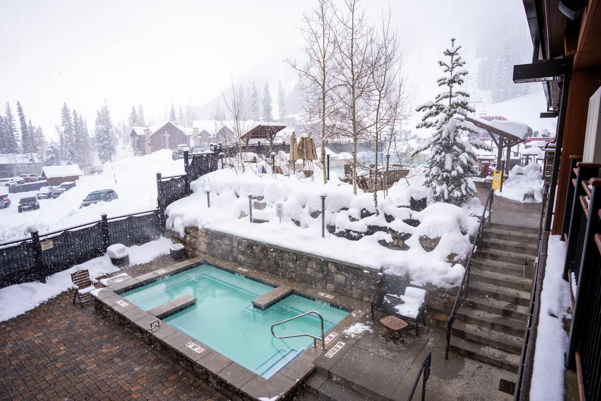An inviting outdoor hot tub surrounded by snow and trees, with steam rising into the cold winter air. The scene is serene, capturing the essence of relaxation after a day on the slopes, and hints at shared moments of warmth and tranquility.