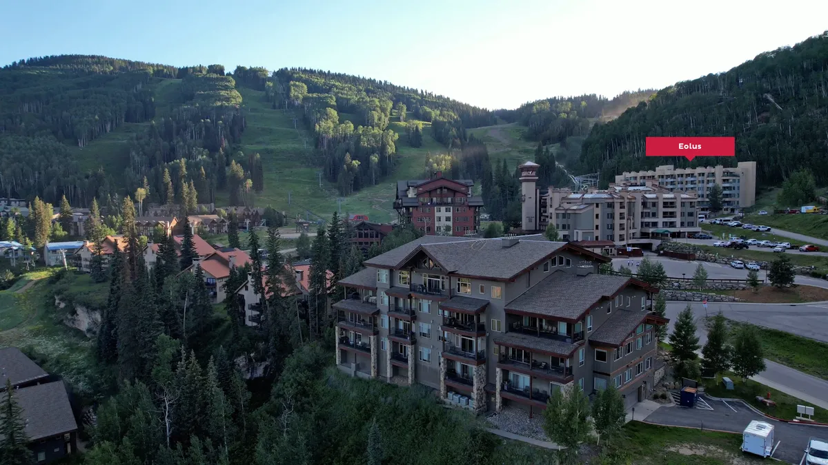 The exterior of the resort, framed by a mountain backdrop and surrounded by trees. The image includes the hotel architecture and a visible parking area, suggesting convenience and ease of access. The mood is adventurous and serene, hinting at the energetic village plaza nearby.