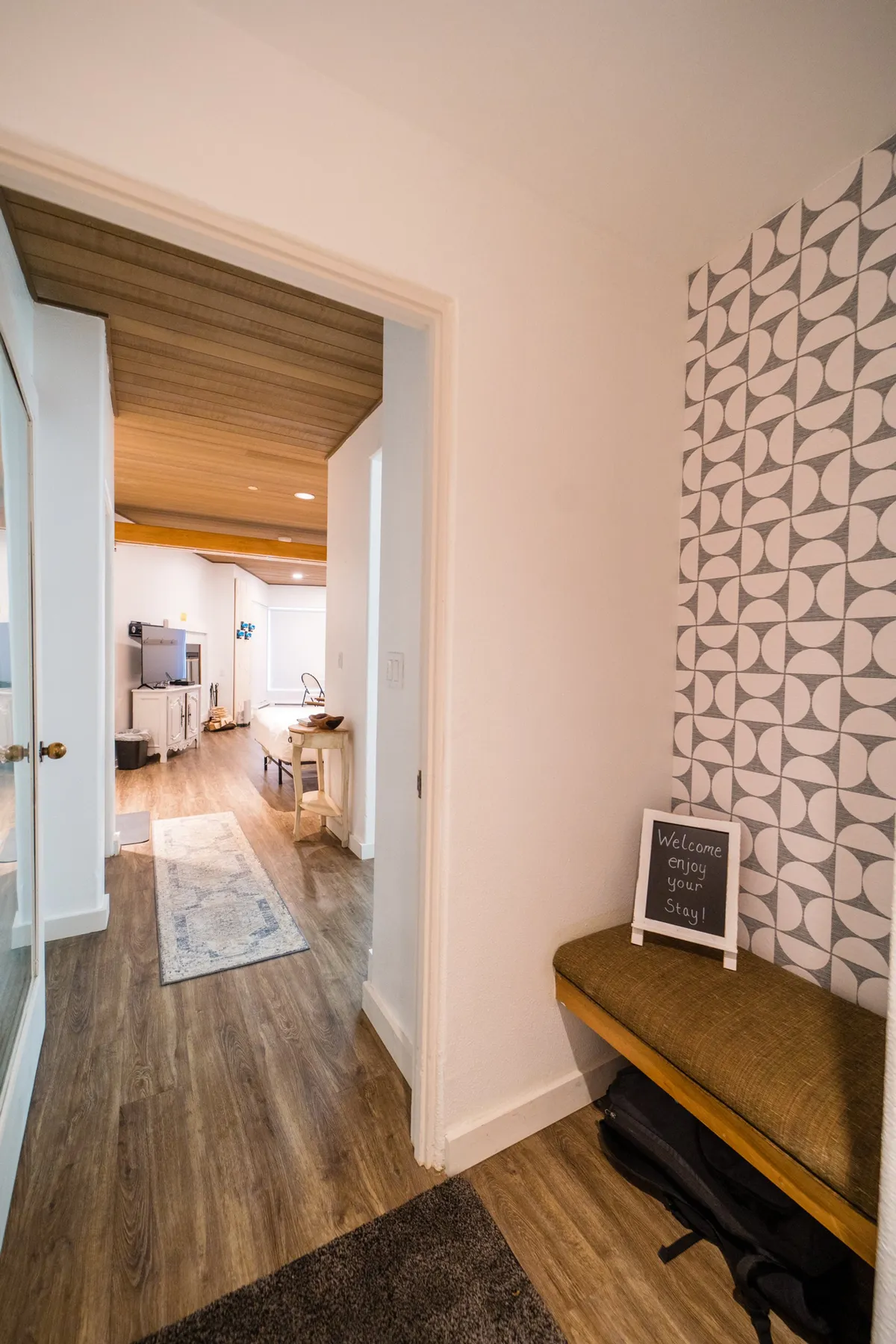 A welcoming entryway with a geometric patterned wall, a friendly 'welcome' sign, and a wooden bench, evoking a sense of hospitality and arrival. The scene suggests the start of an adventure, with guests preparing to step out into the vibrant plaza beyond.