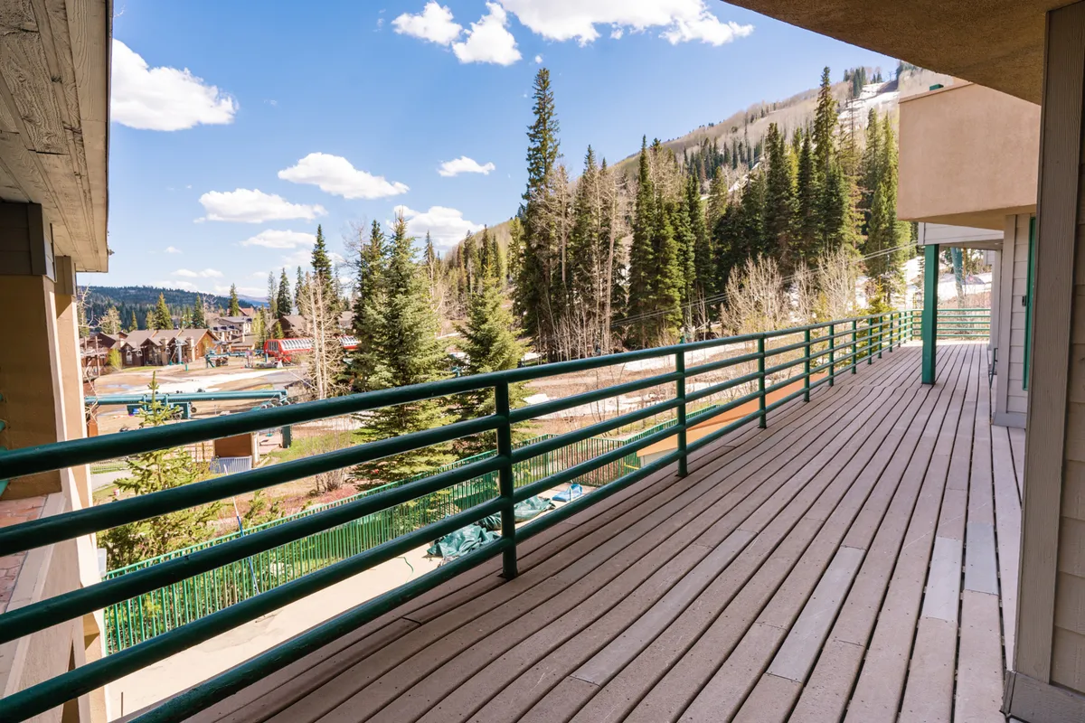 A serene wooden deck bordered by green railings overlooks a sweeping mountain landscape, with surrounding trees framing unobstructed views. The deck is spacious, inviting guests to relax and enjoy nature at any time of day.