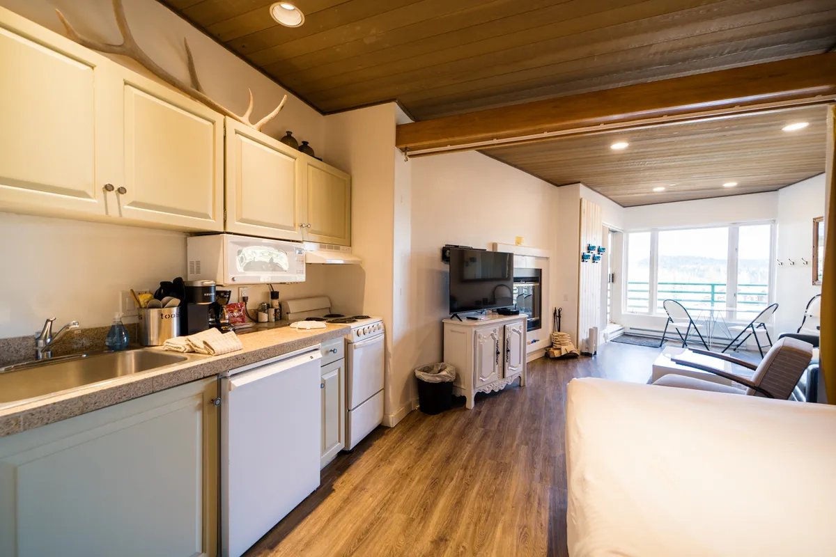 This image showcases an inviting studio living area featuring a kitchen, a cozy living space with a fireplace, large windows that provide panoramic mountain views, and a television. The arrangement highlights the open-plan layout and the blend of comfort and scenic beauty.