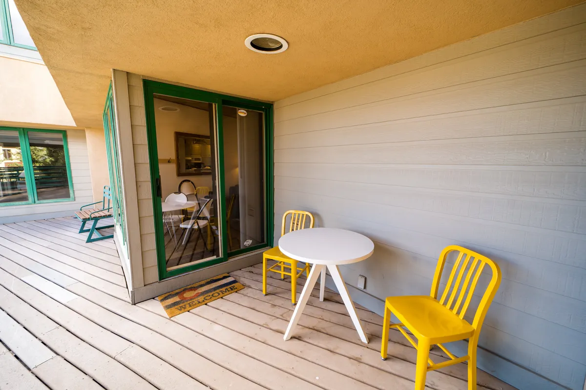 A cheerful patio scene featuring two yellow chairs and a white table outside a green door, with a welcoming mat at the entrance. The setup suggests a quiet, sunny spot perfect for relaxing after outdoor adventures or enjoying a morning coffee while taking in fresh mountain air.