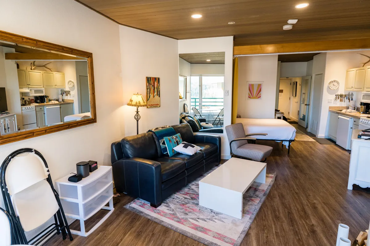 This image shows a comfortable studio-style living area with a sofa, coffee table, decorative artwork, and a kitchen area in the background. The ambiance is relaxed, ideal for socializing or quiet moments. The scene hints at a balcony or outdoor space, tying in with the idea of morning coffee and mountain views.
