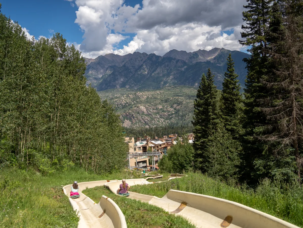 This exterior image captures a lively summer scene with people enjoying the sliding tracks against a dramatic mountain backdrop. Lush green trees and open sky frame the action, conveying a sense of adventure and natural beauty synonymous with the San Juans in summer.
